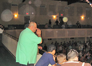 Man in a green shirt speaking into a microphone, with circular light spots (angel orbs) in the photograph